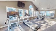 This well-appointed fitness center at 4625 71st St in Lubbock, Texas is designed to provide a bright, motivating environment for a comprehensive workout experience. The space is defined by its dramatic vaulted ceilings with exposed beam accents, which lend an open, airy feel throughout. A striking arched window serves as a focal centerpiece on one wall, flanked by a series of large, multi-pane windows that flood the interior with natural light and offer views of the outdoor pool area, creating an energizing atmosphere during any workout.

The cardio zone is well-equipped with multiple treadmills, elliptical trainers, and a stationary cycle, all positioned to take advantage of the pool-facing windows. Wall-mounted flat-screen televisions are strategically placed for easy viewing from the cardio equipment. A ceiling fan ensures comfortable air circulation throughout the space.

The strength training area features a full rack of dumbbells in graduated weights alongside a multi-function cable machine and a workout bench, accommodating a range of resistance exercises. Glass-panel sliding doors connect this section to an adjoining corridor, providing easy access and an open sightline. The flooring throughout consists of durable, patterned carpet with darker tiling accents in the cardio zone. The neutral taupe wall tones and clean white trim complete the polished, well-maintained aesthetic of this community fitness facility.
