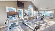 This well-appointed fitness center at 4625 71st St in Lubbock, Texas is designed to provide a bright, motivating environment for a comprehensive workout experience. The space is defined by its dramatic vaulted ceilings with exposed beam accents, which lend an open, airy feel throughout. A striking arched window serves as a focal centerpiece on one wall, flanked by a series of large, multi-pane windows that flood the interior with natural light and offer views of the outdoor pool area, creating an energizing atmosphere during any workout.

The cardio zone is well-equipped with multiple treadmills, elliptical trainers, and a stationary cycle, all positioned to take advantage of the pool-facing windows. Wall-mounted flat-screen televisions are strategically placed for easy viewing from the cardio equipment. A ceiling fan ensures comfortable air circulation throughout the space.

The strength training area features a full rack of dumbbells in graduated weights alongside a multi-function cable machine and a workout bench, accommodating a range of resistance exercises. Glass-panel sliding doors connect this section to an adjoining corridor, providing easy access and an open sightline. The flooring throughout consists of durable, patterned carpet with darker tiling accents in the cardio zone. The neutral taupe wall tones and clean white trim complete the polished, well-maintained aesthetic of this community fitness facility.