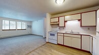 This single-floor, 2-bedroom unit offers a practical and efficiently arranged layout. The open-concept kitchen and living area forms the central hub of the space, with light hardwood-style flooring in the kitchen transitioning to carpet in the living area. The kitchen is equipped with white appliances, including an electric range, refrigerator, and dishwasher, alongside cream cabinetry accented with dark red trim. An under-cabinet light above the range and a double sink add functional detail. The living area benefits from natural light through a bank of windows and is equipped with a wall-mounted air conditioning unit and baseboard heating for year-round climate control. A textured ceiling runs throughout the main living spaces. Both bedrooms feature wall-to-wall carpet, baseboard heating, and sliding-door closets with built-in shelving. The primary bedroom is a comfortable size with a window providing natural light. The second bedroom also includes a wall unit and closet. Overall, the unit presents a clean, neutral palette throughout, creating a straightforward and functional living environment.