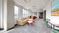 Located at 1184 Hank Aaron Dr in Atlanta, GA, this contemporary office suite offers a well-appointed single-floor layout designed for both collaboration and focused work. The space is defined by large, black-framed windows that wrap around the perimeter, flooding the interior with natural light and providing sweeping views of the surrounding Atlanta cityscape.

The main lounge area serves as the heart of the suite, furnished with a plush sectional sofa, cognac leather accent chairs, and patterned carpeting that adds warmth and texture to the modern aesthetic. This open area flows seamlessly into additional seating zones, making it ideal for informal gatherings or relaxed collaboration.

A dedicated conference room features a substantial dark-topped table surrounded by white upholstered chairs, offering a professional setting for meetings. Glass-paneled doors connect it to the main space, maintaining visual openness while providing acoustic separation. A smaller breakout room with a round marble-top table and wooden chairs offers an intimate setting for focused discussions, accented by decorative wall art.

The suite also includes a private office with glass-panel door access, as well as a built-in kitchenette area with white cabinetry and a countertop for added convenience. Throughout, the clean gray walls, patterned carpet, and carefully curated furnishings create a polished, professional atmosphere.