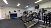 This single-floor fitness center offers a comprehensive workout environment designed to accommodate a wide range of exercise routines. The cardio area is outfitted with multiple treadmills from Vision Fitness and Star Trac, as well as a stair climber, all arranged in a row and complemented by wall-mounted flat-screen televisions for entertainment during workouts. Large mirrors line the walls, enhancing the sense of space and allowing users to monitor their form throughout their sessions. Fluorescent ceiling lighting ensures the entire facility is well illuminated.

The strength and functional training zone features a dumbbell rack with a variety of weights, an adjustable workout bench, and a multi-station cable machine. A dedicated wall-mounted medicine ball rack provides options for dynamic and functional exercises. A recumbent stationary bike offers a low-impact cardio alternative, while additional cable and pulley attachments support upper-body training.

Throughout the space, University of Arizona Wildcats signage and logos reinforce a spirited athletic atmosphere. Dark commercial-grade carpet covers the floor, providing durability underfoot. A standing fan and a wall-mounted sanitation dispenser reflect attention to user comfort and hygiene. The open layout and diverse equipment selection make this fitness center suitable for both cardio-focused and strength-training workout regimens.