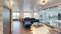 This sophisticated single-floor space is designed with a strong emphasis on modern aesthetics and open, social living. The main lounge area is anchored by rich dark hardwood flooring, tray ceilings with recessed lighting, and a pair of striking sputnik-style gold chandeliers that serve as focal points overhead. Generous natural light pours in through large windows, revealing views of a waterfront beyond, while a wall-mounted flat-screen television and an eclectic mix of tufted blue velvet sofas and neutral accent chairs create an inviting, curated atmosphere. A cowhide area rug and a glass-top coffee table add to the layered, contemporary design.

Floor-to-ceiling glass partitions with metal railings divide the space with architectural elegance, maintaining visual openness while defining separate zones. A game or billiards area is visible in the background, further highlighting the entertainment-focused design.

The wet bar area transitions seamlessly from the lounge and features white quartz countertops, dark cabinetry, a mosaic tile backsplash in cool grey tones, open floating shelves, and stainless steel appliances. A textured stacked stone accent wall adds a striking natural contrast to the otherwise sleek, modern palette. A high-top bistro table with metal stools completes this functional yet stylish refreshment station.