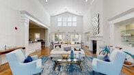 This well-appointed single-story clubhouse space exudes a welcoming, coastal-inspired elegance from the moment you step through its grand entrance. The foyer greets visitors with warm hardwood flooring, soaring double-height ceilings, and an abundance of natural light streaming through large transom windows and glass-paneled doors that frame views of the surrounding greenery.

The central gathering area serves as the heart of the space, featuring dramatic vaulted ceilings that amplify the sense of openness. A classic white fireplace anchors one wall, complemented by a decorative geometric mirror and built-in glass display shelving with integrated lighting. The lounge is furnished with a mix of crisp cream sofas and soft blue accent chairs, all centered around a glass-and-gold coffee table atop a rich blue area rug — creating a cohesive coastal aesthetic.

Multiple seating arrangements and French doors along the rear wall allow for flexible use of the space while maintaining a strong indoor-outdoor connection. A dedicated business center, accessible through wide, column-framed doorways, provides workstations equipped with computers and ample desk space. A wet bar or café counter area is also visible, adding to the functionality of this multi-purpose facility. Throughout, crisp white millwork, polished hardwood floors, and carefully curated décor contribute to a refined yet approachable ambiance.