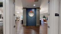 This expansive single-floor space offers a thoughtfully designed layout spanning a substantial footprint, combining social, functional, and utility areas in a cohesive modern aesthetic. The entry corridor features light tile flooring, clean white millwork, and glass-paneled partitions that provide sightlines into adjacent spaces, creating an open and welcoming arrival experience.

The main living area serves as the centerpiece of the space, anchored by a sleek linear fireplace framed in stacked stone tile with a wall-mounted television above. Recessed lighting and tray ceiling details add architectural depth, while hardwood flooring flows throughout the social zones. Bold navy accent walls featuring geometric relief paneling appear in multiple areas, lending a distinctive design character. A circular mirror element echoes this motif in the hallway transition between spaces.

The kitchen is equipped with stainless steel appliances, rich dark cabinetry, granite countertops, and a large central island beneath cylindrical pendant lights. A cozy dining nook with built-in banquette seating, a bronze-toned round table, and a navy geometric accent wall provides an intimate dining option. A formal conference-style dining room with a tray ceiling and chandelier offers a more structured gathering space. Package lockers and a wall-mounted television are conveniently housed within a secondary hallway area.