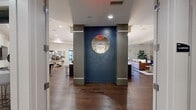 This expansive single-floor space offers a thoughtfully designed layout spanning a substantial footprint, combining social, functional, and utility areas in a cohesive modern aesthetic. The entry corridor features light tile flooring, clean white millwork, and glass-paneled partitions that provide sightlines into adjacent spaces, creating an open and welcoming arrival experience.

The main living area serves as the centerpiece of the space, anchored by a sleek linear fireplace framed in stacked stone tile with a wall-mounted television above. Recessed lighting and tray ceiling details add architectural depth, while hardwood flooring flows throughout the social zones. Bold navy accent walls featuring geometric relief paneling appear in multiple areas, lending a distinctive design character. A circular mirror element echoes this motif in the hallway transition between spaces.

The kitchen is equipped with stainless steel appliances, rich dark cabinetry, granite countertops, and a large central island beneath cylindrical pendant lights. A cozy dining nook with built-in banquette seating, a bronze-toned round table, and a navy geometric accent wall provides an intimate dining option. A formal conference-style dining room with a tray ceiling and chandelier offers a more structured gathering space. Package lockers and a wall-mounted television are conveniently housed within a secondary hallway area.