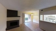 This single-floor space offers a comfortable and functional layout that combines the living and dining areas in an open, connected fashion. The living room is anchored by a classic brick fireplace with a dark wood mantle, above which a large wall-mounted television is positioned — a standout feature that draws the eye upon entry. Soft carpet flooring defines the living area, while hard surface flooring transitions into the dining space, creating a natural visual distinction between the two zones.

The dining area is illuminated by an elegant chandelier and flows seamlessly toward a set of sliding glass doors that open to an outdoor patio, allowing natural light to pour into the interior. The view through the glass doors reveals lush greenery, adding a refreshing outdoor connection to the space.

A pass-through window or counter connects to what appears to be a kitchen area, offering convenient access between spaces while maintaining an open feel. The textured ceiling adds a classic touch throughout, and neutral wall tones keep the atmosphere bright and versatile. HVAC vents are integrated into the ceiling, ensuring year-round comfort. Overall, the space presents a warm, inviting ambiance well-suited for both everyday living and casual entertaining.