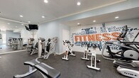 Located at 1525 Station Center Blvd in Suwanee, GA, this fitness center offers a generously sized, single-floor exercise room designed to support a full range of workout routines. The space features durable rubber flooring throughout, providing a safe and practical surface for all types of training. Recessed lighting and dual ceiling fans ensure the room remains bright and comfortable during use.

A bold, typographic fitness-themed mural serves as a striking focal point along one wall, adding energy and motivation to the environment. Large French-style windows and glass doors along one side of the room invite natural light and offer views of the outdoors, enhancing the overall atmosphere.

The facility is comprehensively equipped with a variety of cardio machines, including treadmills, stationary spin bikes, and an elliptical. Strength training is well-represented with multiple selectorized resistance machines, a cable cross machine, and a full rack of dumbbells. Functional fitness accessories are also available, including kettlebells in varying weights, stability balls, medicine balls, and resistance bands. A built-in shelving unit with cabinetry provides organized storage for smaller equipment. Multiple wall-mounted flat-screen televisions are positioned throughout the room for entertainment during workouts, completing this thoughtfully outfitted fitness space.