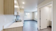 This compact single-floor unit is thoughtfully designed to maximize functionality within a efficient footprint. The open layout connects the kitchen and main living area seamlessly, creating a cohesive and practical space. Neutral cream-toned walls and durable wood-look flooring in a cool gray finish run throughout, contributing to a clean, contemporary aesthetic.

The kitchen is outfitted with light wood-grain cabinetry spanning both upper and lower sections, offering ample storage. Countertops feature a light stone-look surface, complemented by a stainless steel sink with a modern faucet. White appliances, including a refrigerator and over-range microwave, complete the kitchen setup.

The main living area benefits from multiple large windows fitted with horizontal blinds, allowing generous natural light while maintaining privacy. Views of mature trees are visible from the corner windows, adding a pleasant natural element to the space. A wall-mounted radiator provides heating.

The bathroom is cleanly appointed with a pedestal sink, toilet, and shower-tub combination. A mirrored medicine cabinet above the sink adds storage. Wood-look flooring continues here, maintaining visual consistency throughout the unit. Recessed ceiling lighting illuminates the space efficiently. The unit is accessed through a secure entry door with a keypad.
