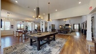 Located at 726 Cumberland Trace Rd in Bowling Green, Kentucky, this single-floor community clubhouse offers a thoughtfully designed gathering space with a variety of distinct functional areas. The interior is anchored by rich hardwood flooring that flows throughout, tying together each zone with a cohesive, upscale aesthetic.

The game room area features a full-size pool table set atop a patterned area rug, illuminated by striking modern pendant chandeliers with brass and black finishes. Adjacent to the game area, casual seating includes a cozy nook with tufted orange leather armchairs positioned beside tall windows that flood the space with natural light.

The lounge area is a standout, boasting soaring coffered ceilings, a dramatic linear fireplace with sleek stone surround, and an assortment of sophisticated seating arrangements. Clerestory windows line the upper walls, drawing in additional daylight and enhancing the sense of openness. Warm-toned French doors provide views and access to the outdoors.

A dining and social area features mid-century modern pub tables and bar seating set against a bold mosaic tile accent wall, with pendant lighting overhead. White decorative columns throughout define spaces without closing them off, maintaining an airy, open feel. The overall ambiance is modern yet welcoming, blending elegance with casual comfort.