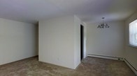This single-floor space presents a straightforward, functional layout with clean lines and a neutral aesthetic throughout. The interior walls are finished in a light off-white tone, creating a bright and open atmosphere that allows for versatile decorating possibilities. A partial wall division separates areas of the space, with a doorway providing access to an adjoining room, suggesting a practical flow between living areas. The flooring appears to be a textured, stone-like material in muted earth tones, adding subtle visual interest underfoot. A multi-arm chandelier with classic styling serves as a focal point in one section of the space, providing overhead lighting and a modest decorative element. Along the base of the wall, a baseboard heating unit is visible, indicating a dedicated heating system for year-round comfort. Electrical outlets are positioned at standard heights along the walls, offering practical utility throughout. The ceilings are smooth and white, contributing to the overall sense of light and openness. Currently unoccupied, the space serves as a clean, neutral canvas well-suited for customization to suit a variety of uses or design preferences.