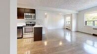 Located on Queen Street East in Toronto's Upper Beaches neighbourhood, this 2-bedroom, 1-bathroom unit offers a well-designed layout with consistent hardwood flooring throughout. The open-concept kitchen and living area forms the heart of the space, where a modern kitchen features rich dark wood cabinetry, stainless steel appliances including a refrigerator, range, and over-the-range microwave, white quartz countertops, and a herringbone-patterned tile backsplash. The kitchen flows seamlessly into a generously sized living area bathed in natural light from large windows overlooking greenery. The primary bedroom is a comfortable size, featuring hardwood floors, a closet, and an adjacent full bathroom. The second bedroom also benefits from hardwood floors and a bright corner window with radiator heating. The full bathroom is finished with neutral beige tile surrounding a soaker tub and includes a window for natural ventilation. Textured ceilings, crisp white trim, and radiator heating units are consistent throughout, lending the space a classic character. The bright, open living area with multiple windows providing views of the surrounding neighbourhood gives the unit an airy and welcoming feel.