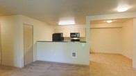 This single-floor residence is designed with an open and functional layout that maximizes the sense of space throughout. The kitchen serves as a central hub, featuring light-toned cabinetry, a dark countertop peninsula that doubles as a breakfast bar or prep space, and black appliances including a refrigerator, microwave, and range. The peninsula design creates a natural divide between the kitchen and the carpeted living area, while maintaining an open, connected feel. Recessed and overhead lighting illuminate the kitchen workspace efficiently. The living area is finished with wall-to-wall carpeting in a neutral tone, contributing to a warm and comfortable ambiance. An open doorway visible from the main living space leads into an additional room, also carpeted, offering flexible use as a bedroom or secondary living area. The walls throughout are finished in a soft, neutral palette, lending a clean and move-in-ready appearance. The overall design emphasizes practicality and ease of movement between spaces, making the layout well-suited for everyday living. The unit is situated entirely on one level, ensuring convenient, step-free access throughout.