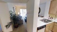 This single-floor residence offers a functional and comfortable layout with a seamless flow between its main living spaces. The living room serves as the heart of the home, featuring soft wall-to-wall carpeting, warm neutral-toned walls, and a large sliding glass door that invites an abundance of natural light while providing a direct view of lush outdoor greenery. A decorative palm plant adds a touch of organic warmth to the space, complementing the relaxed ambiance.

The kitchen is positioned adjacent to the living area and is equipped with white appliances, including a dishwasher, and a countertop sink. Light wood cabinetry lines the kitchen walls, offering ample storage while contributing to the home's warm, neutral aesthetic. A pass-through column between the kitchen and living room creates a sense of openness while maintaining a defined separation between the two spaces.

Throughout the unit, textured ceilings add a classic residential character. The overall design emphasizes practicality and comfort, with a cohesive color palette of beige and white tones that creates a bright, welcoming atmosphere. Natural light flows generously through the space, enhancing the airy feel of the interior.