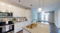 Located at 900 Doverside Dr in Apex, NC, this single-floor residence offers a thoughtfully designed open-concept layout that seamlessly connects the kitchen, dining, and living areas. The kitchen is a standout feature, showcasing crisp white shaker cabinetry, granite countertops, a large center island with a double undermount sink, pendant lighting, and a full suite of stainless steel appliances including a range, microwave, and refrigerator. Rich, dark hardwood-style flooring runs throughout the main living areas, adding warmth and a polished aesthetic. The living area is bathed in natural light, anchored by French doors that open to a private balcony with exterior views, and finished with a ceiling fan overhead. The bedroom is carpeted for comfort, features a ceiling fan, and includes a generous window that fills the room with natural light. The bathroom is well-appointed with granite-topped white vanity cabinetry, tile flooring, a tub-shower combination, and clean, neutral finishes throughout. The overall ambiance of the space is contemporary and inviting, with a consistent soft gray color palette, modern hardware, and an efficient, functional floor plan suited for comfortable everyday living.