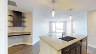This single-floor residence offers a thoughtfully designed open-concept layout that seamlessly connects the kitchen and living areas. The kitchen is a standout feature, highlighted by a spacious granite-topped island with an undermount double sink and dark cabinetry that provides a striking contrast against the light countertops. Elegant pendant lights hang above the island, adding both style and functional illumination. Open shelving along one wall provides convenient storage and display space, complementing the overall contemporary aesthetic.

The kitchen flows naturally into the living area, which is bathed in natural light from large windows that brighten the entire space. A ceiling fan is centrally positioned to enhance comfort throughout the room. Rich dark hardwood flooring runs throughout, adding warmth and continuity between spaces.

The neutral color palette on the walls enhances the sense of openness, while the clean lines and modern finishes give the space a fresh, well-appointed feel. The combination of recessed lighting and pendant fixtures ensures the interior remains well-lit at any hour. Overall, this residence balances practical functionality with modern design, creating an inviting and comfortable living environment.