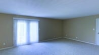 This single-floor space offers a clean, straightforward layout designed for comfortable everyday living. The main living area is a standout feature, characterized by its generous proportions and an abundance of natural light streaming in through a large sliding glass door fitted with white vertical blinds. The door serves as both a functional entry point and a bright focal element that enhances the openness of the room. The living area is finished with plush wall-to-wall carpeting in a neutral tone, providing a warm and inviting underfoot feel. The walls are painted in a soft, earthy beige that complements the light palette throughout the space. White baseboards provide a clean contrast along the perimeter of the room, adding a crisp, finished look to the interior. The ceiling is smooth and generously proportioned, contributing to an airy sense of volume. Electrical outlets are conveniently positioned along multiple walls, offering practical flexibility for furniture arrangement. Overall, the space presents a neutral, move-in-ready canvas that is well-lit, simply appointed, and adaptable to a variety of interior styles and functional needs.