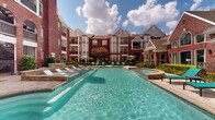 Located at 8787 Woodway Dr in Houston, Texas, this outdoor amenity space is the centerpiece of a well-appointed residential community defined by classic red brick architecture with gabled rooflines, dormer windows, and private balconies. The highlight of the space is a generously sized resort-style swimming pool with crystal-clear turquoise water, surrounded by a travertine stone deck furnished with lounge chairs and poolside seating in vibrant teal accents. Adjacent to the pool, a raised spa provides an additional relaxation option with easy access from the main deck. A standout architectural feature is the large pergola structure, which casts dramatic latticed shadows across the stone patio below and shelters a built-in outdoor fireplace crafted from natural stacked stone — creating an inviting gathering area with Adirondack chairs arranged for casual outdoor entertaining. A built-in grilling station with stone surround adds functional appeal for outdoor dining. Manicured landscaping with lush greenery and ornamental iron fencing frames the entire outdoor courtyard, contributing to a polished, resort-like atmosphere. The multi-story residential buildings enclosing the courtyard provide a sense of privacy and enclosure while overlooking the amenity area, completing a cohesive and well-designed outdoor living environment ideal for relaxation and social gatherings.