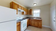 This single-floor space includes a well-organized kitchen that serves as a central feature. The kitchen is outfitted with warm-toned oak cabinetry, offering generous upper and lower storage throughout. Dark laminate countertops provide a contrasting work surface, while a double-basin sink is positioned beneath a window that allows natural light to brighten the room. White appliances, including a gas range with a ventilation hood above and a full-size refrigerator, complete the functional layout. The L-shaped configuration makes efficient use of the available space, providing a practical workflow between the cooking and prep areas. Light ceramic tile flooring runs throughout the kitchen, contributing to a clean, easy-to-maintain finish. A flush-mount ceiling fixture provides overhead lighting. The neutral wall color keeps the atmosphere open and versatile, complementing the natural wood tones of the cabinetry. Overall, the kitchen presents a straightforward, functional design suited for everyday use.