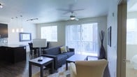 This single-floor space offers a well-designed, open-concept layout that maximizes both comfort and functionality. The living area serves as the heart of the space, bathed in natural light streaming through a large sliding glass door fitted with vertical blinds and a secondary window with horizontal blinds. A ceiling fan with an integrated light fixture anchors the room, providing both airflow and overhead illumination.

The living area is thoughtfully arranged with a contemporary dark sofa accented by decorative pillows, a rectangular coffee table, and a plush accent chair resting atop a patterned area rug that adds visual warmth to the space. A sleek side table with a modern lamp adds ambient lighting and a sense of layered design.

Adjoining the living area is a dedicated dining space, visible in the background, featuring a dark dining table and chairs that complement the overall modern aesthetic. The neutral wall tones and textured ceiling provide a clean, understated backdrop that ties the entire space together. A tall decorative vase near the sliding door adds an artistic finishing touch. Overall, the space presents a bright, airy, and cohesive environment suited for both everyday living and entertaining.