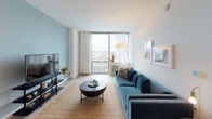 Located on an upper floor at 770 Jackson Ave in Hoboken, NJ, this well-appointed 1-bedroom, 1-bathroom residence combines contemporary design with functional living. The open-concept layout is anchored by a bright living area featuring large floor-to-ceiling windows that flood the space with natural light and offer expansive views of the Hoboken skyline and surrounding cityscape. The living room provides a comfortable and airy atmosphere, with light wood-toned flooring running throughout. The kitchen is outfitted with stainless steel appliances, including a full-size refrigerator, microwave, and range, complemented by rich dark wood cabinetry and sleek white upper cabinets that extend to the ceiling for ample storage. A tiled backsplash adds a refined finishing touch. The bedroom is a serene retreat, featuring clean white walls and access to the same generous natural light found throughout the home. The bathroom is thoughtfully designed with a floating dark wood vanity, white countertop, a large mirror, and a separate walk-in shower, all finished with modern tile and fixtures. Soft blue accent walls appear throughout the residence, tying together a cohesive and calming color palette that enhances the overall modern aesthetic of this Hoboken home.