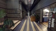 This space combines a grand building lobby with an adjoining coffee shop, each offering a distinct yet complementary atmosphere. The lobby showcases strong Art Deco influences, characterized by dark marble-clad walls, a textured geometric ceiling, and boldly patterned terrazzo flooring with cream and charcoal striping. A central reception desk with task lighting anchors the corridor, flanked by elevator banks and a vintage U.S. mail letterbox mounted near a marble staircase with chrome railings. Potted tropical plants punctuate the space, softening the industrial elegance. The entry vestibule features glass doors that invite natural light, with a lounge sofa and large-leafed plants creating a welcoming transition.

The coffee shop presents a markedly different character—intimate and richly layered. Warm wall sconces and pendant lighting cast a soft glow over a curated mix of furnishings, including a tufted leather sofa, upholstered armchairs, and marble-topped side tables arranged on a patterned area rug. Hanging planters cascade greenery from above, while framed art lines the upper walls. A communal table near the street-facing window offers views to the exterior, and a bar-height counter with stools provides additional seating. Polished concrete flooring and dark wainscoting complete the cozy, eclectic aesthetic.