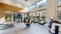 This well-appointed fitness center offers a generous, open layout across a single floor, designed to accommodate a full range of workout activities. The space is defined by soaring double-height ceilings that create an airy, energizing atmosphere, complemented by expansive floor-to-ceiling windows that flood the interior with natural light and provide views of the surrounding outdoor courtyard and greenery.

The main workout area is equipped with a comprehensive selection of cardio machines, including treadmills, ellipticals, and stationary bikes, all arranged along the windowed wall to maximize natural light and views. Strength training is well represented with a multi-functional cable and weight machine station, a full dumbbell rack, and additional resistance equipment. Herringbone-patterned flooring runs throughout, adding a refined aesthetic detail to the space.

A dedicated studio room, separated by sleek glass partition walls and doors, provides a versatile space for cycling classes and functional training, outfitted with spin bikes, TRX suspension systems, and a large wall-mounted display screen. Floor-to-ceiling mirrors enhance the sense of space within the studio.

Distinctive design elements include warm wood paneling accents, a striking living moss wall installation that adds a natural, biophilic element, and recessed lighting throughout. The overall ambiance is clean, modern, and motivating.