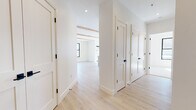 Located at 82–98 Jolley Drive in Bloomfield, Connecticut, this newly constructed single-floor residence showcases a clean, contemporary design defined by a bright, neutral palette and high-quality finishes. Light hardwood flooring runs throughout the main living areas and hallways, while exposed natural wood ceiling beams add warmth and architectural character to the open-concept living and kitchen spaces.

The kitchen is a standout feature, equipped with flat-front light wood cabinetry extending to the ceiling, stainless steel appliances including a refrigerator, microwave, and range, and a large island with a white quartz countertop and an integrated sink fitted with a matte navy faucet. A modern globe pendant light fixture and recessed lighting complete the space. Black-framed windows allow generous natural light to flow in and offer views of the surrounding landscape.

The hallway connects multiple rooms via Shaker-style interior doors with matte black hardware. Bedrooms are finished with plush neutral carpeting and black-framed double windows. Bathrooms are thoughtfully appointed: one features dark veined marble-look tile flooring, a glass-enclosed walk-in shower with white subway tile, and a light wood vanity, while a second bath offers large-format light tile flooring and a matching wood vanity. The entry foyer provides a welcoming transition into the space with its clean lines and hardwood floors.
