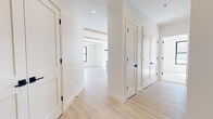 Located at 82–98 Jolley Drive in Bloomfield, Connecticut, this newly constructed single-floor residence showcases a clean, contemporary design defined by a bright, neutral palette and high-quality finishes. Light hardwood flooring runs throughout the main living areas and hallways, while exposed natural wood ceiling beams add warmth and architectural character to the open-concept living and kitchen spaces.

The kitchen is a standout feature, equipped with flat-front light wood cabinetry extending to the ceiling, stainless steel appliances including a refrigerator, microwave, and range, and a large island with a white quartz countertop and an integrated sink fitted with a matte navy faucet. A modern globe pendant light fixture and recessed lighting complete the space. Black-framed windows allow generous natural light to flow in and offer views of the surrounding landscape.

The hallway connects multiple rooms via Shaker-style interior doors with matte black hardware. Bedrooms are finished with plush neutral carpeting and black-framed double windows. Bathrooms are thoughtfully appointed: one features dark veined marble-look tile flooring, a glass-enclosed walk-in shower with white subway tile, and a light wood vanity, while a second bath offers large-format light tile flooring and a matching wood vanity. The entry foyer provides a welcoming transition into the space with its clean lines and hardwood floors.
