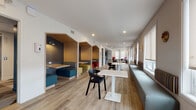 This thoughtfully designed single-floor communal space offers a versatile layout that seamlessly blends focused work, casual dining, and relaxed lounging. The elongated floor plan is anchored by light wood-tone flooring throughout, creating a warm and cohesive aesthetic across all zones.

Along one side of the main corridor, a row of semi-private booth nooks features warm wood paneling, textured dark accent walls, and built-in bench seating with colorful upholstered cushions in teal, yellow, and blue — ideal for focused individual work or small group collaboration. Opposite the booths, a continuous gray banquette runs beneath a generous stretch of windows fitted with roller shades, paired with white pedestal tables and modern black chairs, forming a bright, open dining and work area bathed in natural light.

At one end of the space, a cozy lounge nook offers a more relaxed atmosphere, furnished with vibrant red accent chairs, a gray sofa, a round coffee table, and a soft area rug — all framed by large windows with outdoor views. Recessed ceiling lighting and colorful wall art add warmth and personality throughout.

The space also includes an accessible restroom finished with white subway tile walls, dark slate-toned floor tiles, and clean, functional fixtures. Overall, the space balances a contemporary, welcoming design with practical, multi-use functionality.