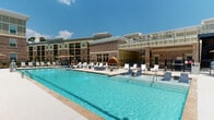 Located at 380 Topwater Dr in Clayton, NC, this outdoor amenity area is designed to deliver a resort-caliber experience. At the heart of the space is a large, sparkling swimming pool with crystal-clear blue water, accessible via stainless steel entry rails and surrounded by a generous concrete sundeck. The deck is well-furnished with rows of chaise lounges, complemented by distinctive pod-style hanging cabana chairs that provide shaded, private seating nooks. Umbrella-shaded seating clusters are distributed throughout the pool deck, offering additional options for relaxation under the North Carolina sun. The space is enclosed by a black iron fence, ensuring safety and a sense of enclosure. Adjacent to the pool area is a covered outdoor pavilion that serves as a sheltered lounge. This semi-open structure features paneled walls in a muted sage-green palette with crisp white trim detailing, ceiling fans for airflow, and an outdoor television. The pavilion is furnished with large sectional sofas in bold blue upholstery, Adirondack-style chairs, and round tables, creating a comfortable gathering space. A built-in outdoor kitchen or grill area is also visible within the pavilion. The surrounding multi-story brick and siding building architecture frames the courtyard, lending a cohesive, upscale residential aesthetic to the entire outdoor space.