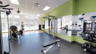 This fitness center is thoughtfully designed to support a well-rounded workout experience. The space features a drop-ceiling with recessed panel lighting that ensures consistent, even illumination throughout. A row of large windows along one wall floods the room with natural light while offering views of the outdoor pool area, creating an energizing atmosphere for exercise.

The flooring transitions between rubberized exercise matting and hardwood-style surfaces, providing appropriate traction and support for a variety of workout activities. A bold green accent wall serves as a visual anchor and backdrop for a central multi-functional training rig, which includes TRX suspension trainers and ladder attachments for functional fitness and bodyweight exercises. Floor-to-ceiling mirrors are mounted along one wall, allowing users to monitor their form during workouts.

Additional equipment includes an adjustable weight bench, medicine balls, and cardio machines visible in the mirrored reflection. A sound-absorbing panel is mounted near the windows, contributing to the overall comfort of the space. The layout is open and uncluttered, allowing ample room for stretching, free movement, and equipment use simultaneously.