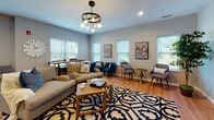 Located at 3 Rindle Rd in Boylston, Massachusetts, this single-floor community amenity space offers a well-appointed combination of social and recreational areas designed for comfort and activity. The lounge area is bright and inviting, featuring warm hardwood flooring, large windows fitted with blinds that allow generous natural light to fill the room. Thoughtfully furnished with a plush sofa, teal accent chairs, and coordinating side seating arranged around a bold patterned area rug, the space is anchored by a stylish multi-bulb chandelier overhead. Framed artwork and a narrow console table along the windowed wall add character and charm. Adjacent to the lounge, the fitness center is spacious and well-equipped with rubber flooring, ceiling fans, and large mirrors that enhance the sense of space. It features cardio machines including ellipticals, a stationary bike, and a stair climber, alongside a cable machine, weight bench, a full rack of dumbbells, and an extensive selection of resistance bands and exercise balls organized on a dedicated rack. A wall-mounted flat-screen TV completes the workout environment. A compact coffee bar area rounds out the amenities, showcasing dark granite countertops, white cabinetry, floating corner shelves, patterned tile flooring, and a built-in beverage refrigerator — perfect for a quick refreshment.
