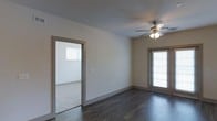 This single-floor residence offers a straightforward and functional layout designed with clean, modern finishes throughout. The main living area features rich, dark hardwood flooring that contrasts elegantly with the neutral, light-toned walls, creating a warm yet contemporary aesthetic. A ceiling fan with an integrated light fixture is centrally positioned overhead, providing both air circulation and ambient lighting to the space. Natural light flows in through a window fitted with blinds, brightening the room and enhancing its open feel. An open doorway connects the main living area to an adjoining room, which features plush carpeting and its own window — offering a softer, cozier atmosphere well-suited for use as a bedroom or additional living space. Consistent trim and neutral wall tones carry throughout both rooms, contributing to a cohesive and unified design. The layout is practical and accessible, making efficient use of the available square footage across a single level. The overall ambiance is clean and move-in ready, with a neutral palette that provides a versatile backdrop for a variety of interior styles and furnishings.