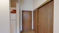 This single-floor unit offers a practical and efficiently organized layout across a modest footprint, comprising a hallway, kitchen, bedroom, and bathroom. The entry hallway greets residents with notably tall ceilings that lend a sense of openness throughout the unit, alongside a large sliding wood-panel closet that provides generous built-in storage from the moment you step inside.

The kitchen features warm oak cabinetry extending to the upper walls, maximizing vertical storage space. Light-toned countertops with a double stainless steel sink and wood-look flooring complete the functional cooking area.

The bedroom is a comfortably sized, carpeted room with high ceilings and a window that brings in natural light. A wide sliding-door closet with built-in wire shelving and hanging rods offers substantial storage capacity. Baseboard heating runs along the exterior wall, ensuring efficient climate control.

The bathroom is designed with accessibility in mind, featuring a full tub-shower combination equipped with a handheld showerhead, multiple grab bars, and a wall-mounted vanity mirror with overhead lighting. The overall ambiance of the unit is clean and neutral, with white walls and light carpeting providing a simple, easy-to-personalize canvas.