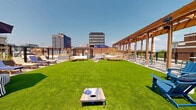 Located at 808 Chapel Street in the heart of New Haven, Connecticut, this expansive rooftop terrace offers a generously sized outdoor retreat with a compelling mix of design elements and recreational amenities. The centerpiece of the space is a large, vibrant artificial turf lawn that provides a versatile area for lounging, lawn games such as cornhole, and casual gatherings. Flanking one side of the terrace is a striking wood pergola structure outfitted with overhead speakers and lighting, creating a shaded corridor with teak deck tile flooring that adds warmth and texture underfoot. The pergola frames sweeping, unobstructed views of the New Haven skyline, including neighboring mid-rise office and residential buildings. Throughout the terrace, a variety of seating arrangements can be found, including modular sectional sofas, Adirondack chairs, striped bean bag chairs, sun loungers, and a hammock boat, all thoughtfully positioned to encourage relaxation. Patio umbrellas provide additional shade across both the turfed and tiled zones. A checkerboard-patterned hardscape area near the pergola adds a playful design detail, while planters with ornamental grasses introduce organic texture. The rooftop is enclosed by black metal railings that ensure safety without obstructing the panoramic urban vistas, making this an exceptional outdoor amenity space.
