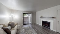 This single-floor residence offers a thoughtfully designed layout with modern updates throughout. The living room serves as the heart of the space, showcasing wood-style laminate flooring, a classic white-surround fireplace with a rustic wood mantle, and a textured ceiling. A sliding glass door floods the room with natural light and opens to a private balcony, offering pleasant outdoor views. A ceiling fan enhances comfort in the adjoining area, which connects seamlessly to the rest of the home via a well-proportioned hallway with consistent flooring throughout. The kitchen is a standout feature, outfitted with crisp white shaker-style cabinetry, granite-look countertops, and white appliances including a refrigerator, range, dishwasher, and over-the-range microwave — all set atop light wood-style flooring that brightens the space. The bathroom continues the home's cohesive aesthetic with matching white cabinetry, granite-look countertops, and neutral tile-style flooring. The bedroom is a calm retreat, featuring the same wood-style flooring, neutral warm-toned walls, and a window that frames a view of surrounding greenery, bringing in soft natural light. Throughout the home, a consistent neutral color palette and updated finishes create a clean, modern ambiance.