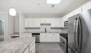 This 990-square-foot, single-floor apartment — Design 2A Premier #091 — offers 2 bedrooms and 2 bathrooms in a well-appointed layout at 141 Park at N Hills St in Raleigh, NC. The open-concept kitchen and living area serves as the heart of the home, featuring rich wood-look luxury vinyl plank flooring, crown molding, and ceiling fans. The kitchen is outfitted with white shaker-style cabinetry, granite countertops, a subway tile backsplash, stainless steel appliances including a refrigerator, microwave, electric range, and dishwasher, and a curved peninsula ideal for casual dining. A built-in desk nook adds functional workspace. The living room is spacious and bright, with crown molding and an arched hallway leading to the bedrooms. Both bedrooms feature plush carpeting, crown molding, and natural light from windows with blinds. The primary bedroom measures 170 square feet and includes a ceiling fan and dedicated closet. Each bedroom has its own closet for added storage. The two bathrooms feature wood-look LVP flooring and white cabinetry with neutral countertops — one with a tub/shower combo and one with a glass-enclosed walk-in shower.