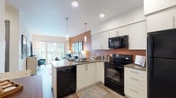 This thoughtfully designed single-floor residence offers a seamless blend of modern aesthetics and functional living. The open-concept kitchen is outfitted with crisp white cabinetry, dark quartz countertops, a suite of black appliances including a refrigerator, electric range, built-in microwave, and dishwasher, and a peninsula with pendant lighting that doubles as a casual dining area. The kitchen flows naturally into the living space, which is bathed in natural light and features warm wood-tone flooring throughout. The living room is anchored by large sliding glass doors that open onto a private balcony, offering views of the surrounding urban landscape. The bedroom is a restful retreat highlighted by a bold blue accent wall, large floor-to-ceiling windows that invite abundant natural light, and generous proportions for comfortable furnishings. The full bathroom is finished with white cabinetry, dark countertops, a wide vanity mirror, and tile flooring arranged in a herringbone pattern, providing both style and practicality. Recessed lighting and clean wall finishes contribute to the bright, contemporary ambiance found throughout the entire residence.