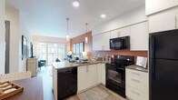 This thoughtfully designed single-floor residence offers a seamless blend of modern aesthetics and functional living. The open-concept kitchen is outfitted with crisp white cabinetry, dark quartz countertops, a suite of black appliances including a refrigerator, electric range, built-in microwave, and dishwasher, and a peninsula with pendant lighting that doubles as a casual dining area. The kitchen flows naturally into the living space, which is bathed in natural light and features warm wood-tone flooring throughout. The living room is anchored by large sliding glass doors that open onto a private balcony, offering views of the surrounding urban landscape. The bedroom is a restful retreat highlighted by a bold blue accent wall, large floor-to-ceiling windows that invite abundant natural light, and generous proportions for comfortable furnishings. The full bathroom is finished with white cabinetry, dark countertops, a wide vanity mirror, and tile flooring arranged in a herringbone pattern, providing both style and practicality. Recessed lighting and clean wall finishes contribute to the bright, contemporary ambiance found throughout the entire residence.