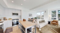 This well-appointed 2-bedroom, 2-bathroom residence is designed with a clean, modern aesthetic across a single floor. The open-concept kitchen and living area forms the heart of the home, featuring a bright white palette, light hardwood flooring, and large windows that frame lush tree canopy views. The kitchen is fully equipped with stainless steel appliances including a gas range, built-in microwave, and French door refrigerator, complemented by tall shaker-style cabinetry, a herringbone tile backsplash, white quartz countertops, and a freestanding butcher block island with bar seating. The living area is bathed in natural light from a wide bank of windows, creating an airy, welcoming atmosphere. The primary bedroom offers generous natural light through dual windows with garden views, while the second bedroom features its own arched mirror accent and closet access. A dedicated walk-in closet provides ample storage. Both bathrooms are finished in a minimalist style with white floating vanities, large mirrors, and wood-toned flooring. One bathroom includes a patterned tile accent rug area, adding a subtle design detail. Hallways connect each space efficiently, maintaining a cohesive and uncluttered flow throughout the residence.