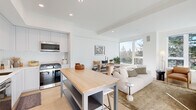 This well-appointed 2-bedroom, 2-bathroom residence is designed with a clean, modern aesthetic across a single floor. The open-concept kitchen and living area forms the heart of the home, featuring a bright white palette, light hardwood flooring, and large windows that frame lush tree canopy views. The kitchen is fully equipped with stainless steel appliances including a gas range, built-in microwave, and French door refrigerator, complemented by tall shaker-style cabinetry, a herringbone tile backsplash, white quartz countertops, and a freestanding butcher block island with bar seating. The living area is bathed in natural light from a wide bank of windows, creating an airy, welcoming atmosphere. The primary bedroom offers generous natural light through dual windows with garden views, while the second bedroom features its own arched mirror accent and closet access. A dedicated walk-in closet provides ample storage. Both bathrooms are finished in a minimalist style with white floating vanities, large mirrors, and wood-toned flooring. One bathroom includes a patterned tile accent rug area, adding a subtle design detail. Hallways connect each space efficiently, maintaining a cohesive and uncluttered flow throughout the residence.