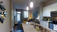 Located on 9th Street in Durham, NC, this single-floor residence showcases a contemporary open-concept design that integrates the kitchen and living areas into one cohesive, functional space. The kitchen is anchored by a generously sized granite countertop island that serves as both a prep surface and a casual gathering point. White cabinetry, a stainless steel sink, and a sleek modern faucet complete the kitchen's clean, polished aesthetic.

Pendant lighting hangs from the ceiling above the island, providing focused illumination that complements the warm ambient lighting found throughout the living area. The living space features dark hardwood flooring grounded by a deep blue area rug, creating a rich contrast against the light gray walls. A large sectional sofa and accent chair are arranged to encourage relaxed conversation and entertainment.

Natural light filters in through multiple windows dressed with layered curtains and blinds, offering both brightness and privacy. A private entry door at the rear of the living area adds convenience and accessibility. The overall ambiance is modern and inviting, with a carefully curated palette of cool grays, navy blues, and warm accent tones throughout.