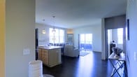 This single-floor space features an open-concept layout that seamlessly connects the kitchen and living areas, creating a cohesive and functional environment. The kitchen is equipped with light wood cabinetry, a peninsula with countertop workspace, and a stainless steel sink. Pendant lights hang above the peninsula, providing focused task lighting while adding a modern design element. The living area accommodates comfortable seating and is anchored by dark hardwood flooring that runs throughout, offering a rich contrast to the bright white walls and ceiling. Large windows along the perimeter of the space allow abundant natural light to fill the interior, contributing to an airy and welcoming ambiance. A sliding glass door provides direct access to an outdoor balcony, extending the living space and bringing in additional light and views of the exterior. The open layout and clean lines throughout give the space a contemporary feel, while the neutral color palette ensures versatility and a sense of openness across the entire floor plan.