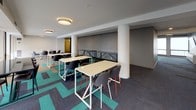 This single-floor multipurpose space is designed to accommodate a range of gathering, meeting, and event needs with a high degree of flexibility. The layout is divided into distinct functional zones, each furnished to support different configurations — from collaborative group work to larger seated assemblies.

The main area features clean-lined white tables paired with contemporary dark chairs, arranged in clusters ideal for small group discussions or workshops. Structural columns punctuate the space, adding an architectural rhythm while maintaining an open, airy feel. Folding tables and stackable chairs are also available, allowing the room to be quickly reconfigured for larger events or presentations. Wall-mounted flat-screen televisions are positioned throughout to support presentations and media display.

A separate, windowed area visible in the layout provides an additional zone with its own seating, benefiting from natural light that filters in through a continuous bank of windows along the exterior wall.

Flush drum pendant lighting is installed throughout, casting a warm, even glow across the space. The standout design element is the boldly patterned carpet — a geometric arrangement of teal, tan, navy, and charcoal tones that energizes the environment and delineates different zones within the open floor plan. Neutral gray walls provide a clean, professional backdrop that keeps the focus on functionality and adaptability.