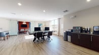 This contemporary single-floor business center is thoughtfully designed to support both focused work and collaborative gatherings. The space features light wood-tone flooring throughout, recessed lighting, and large windows that invite an abundance of natural light and outdoor views, creating a bright and welcoming atmosphere.

The main workstation area is anchored by a sleek, dark-toned communal desk accommodating multiple computer workstations, each equipped with monitors and ergonomic rolling chairs. A beverage and snack station is conveniently positioned nearby, offering easy access to refreshments. An additional dedicated computer nook along the windowed wall provides a more intimate workspace bathed in natural light.

A standout feature of the space is the glass-paneled sliding barn door that opens into a private conference room. The conference room is distinctively styled with bold blue and red accent walls, a wall-mounted flat-screen display, and seating for several attendees around a central table — making it well-suited for meetings and presentations.

Adjacent to the workstation area, a comfortable lounge features a built-in navy blue upholstered banquette set against a textured wave-patterned accent wall. Round marble-top tables paired with contemporary teal and wood-accented chairs, illuminated by globe pendant lights, complete this inviting gathering space.