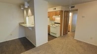 This single-floor residence offers a practical and comfortable layout designed for everyday living. The kitchen serves as a central feature of the home, equipped with warm honey-toned wood cabinetry that provides ample storage space, including a tall pantry-style cabinet for additional organization. A white electric range with a built-in ventilation hood anchors the cooking area, complemented by under-cabinet lighting that adds a warm, functional glow. A peninsula-style breakfast bar with a light-colored countertop extends from the kitchen, creating a casual dining or workspace that also acts as a natural divider between the kitchen and the adjacent living area. The flooring transitions from dark hardwood-style material in the kitchen zone to plush carpeting in the living space, adding texture and warmth to the overall ambiance. The open sightlines between the kitchen, dining, and living areas create a connected, airy feel throughout the home. Neutral wall tones and recessed-style pendant lighting contribute to a clean, understated aesthetic. A hallway visible from the main space suggests additional rooms beyond, offering a sense of depth and functionality to this well-organized single-level home.