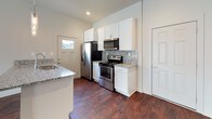 This well-appointed single-floor unit showcases a thoughtfully designed open-concept layout that seamlessly connects the living area to the kitchen. The living space is anchored by dramatic vaulted ceilings that create an airy, expansive atmosphere, complemented by light-toned luxury vinyl plank flooring that runs throughout. A ceiling fan with recessed lighting adds both function and style to the space. Natural light filters in through multiple windows dressed with blinds, brightening the neutral gray and white palette.

The kitchen is outfitted with crisp white shaker-style cabinetry, granite countertops with a mosaic tile backsplash, and a full suite of stainless steel appliances including a range, over-the-range microwave, and refrigerator. A granite-topped breakfast bar with pendant lighting serves as a functional divider between the kitchen and living areas.

The bedrooms continue the consistent flooring and neutral tones, each featuring ceiling fans and natural light from windows. The bathroom is finished with striking floor-to-ceiling vertically laid tile in the shower and tub surround, coordinating marble-look tile flooring, and a granite vanity countertop with white cabinetry — delivering a clean, contemporary aesthetic throughout.