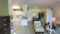 This single-floor residence offers a thoughtfully designed open-concept layout that brings together the kitchen, dining, and living areas in one cohesive, flowing space. The kitchen is outfitted with white cabinetry, white appliances, and a dark-toned countertop that extends into a peninsula, providing both prep space and a natural visual divider between the kitchen and the rest of the living area. Crown molding along the upper cabinetry adds a refined architectural touch. The dining area sits adjacent to the kitchen and features a dark wood table with matching chairs, complemented by decorative wall art and a tall indoor plant that adds a lush, natural element to the room. The living area is carpeted and furnished with a cream upholstered accent chair, creating a warm and inviting atmosphere. Neutral warm-toned walls throughout the space lend a cohesive, calming ambiance. Recessed lighting and flush-mounted fixtures provide ample illumination, enhancing the open and airy feel of the interior. The overall design balances functionality with comfortable, understated elegance.