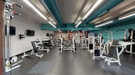 Located at 2 Clocktower in Nashua, NH, this fitness center occupies a single-floor space with a distinctive industrial character. The facility is divided into several clearly defined workout zones, each designed to support a range of exercise disciplines.

The strength training area features durable rubber flooring and an extensive selection of weight machines, free weights, barbells, dumbbells, and benches. Floor-to-ceiling mirrors line the walls, creating a spacious feel and allowing users to monitor their form. Wall-mounted televisions add an entertainment element to the workout experience. Teal-painted exposed ductwork runs along the ceilings, giving the space a modern industrial aesthetic.

The cardio zone offers multiple treadmills, elliptical trainers, and stair climbers set against a backdrop of original exposed brick walls and natural light from traditional multi-pane windows. A separate cycling area houses a row of stationary bikes on carpeted flooring, framed by salmon-colored structural columns with teal accents.

A dedicated free weights room provides a focused environment with mirrored walls, ballet-style bars, and additional dumbbell racks. The facility's entry area features teal-trimmed glass doors and windows, providing a clean transition into the workout spaces. Throughout, the combination of brick, teal, and salmon tones gives the gym a vibrant and energetic ambiance.