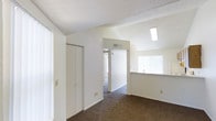 This single-floor residence offers a clean, functional layout designed for comfortable everyday living. The main living area is finished with warm, wall-to-wall carpeting that extends throughout, creating a cohesive and inviting atmosphere. Crisp white walls and a textured ceiling contribute to a bright, airy feel that is further enhanced by natural light streaming in through multiple windows fitted with vertical blinds.

A hallway connects the main living space to additional rooms, providing a practical flow throughout the home. A built-in closet with double doors offers generous storage directly within the living area. The kitchen features a pass-through counter that opens toward the living space, allowing for easy interaction between the two areas. A window above the counter area brings in additional natural light, keeping the kitchen feeling open and pleasant.

An overhead skylight or recessed ceiling fixture adds a further dimension of brightness to the space, ensuring the interior remains well-lit throughout the day. The HVAC vent integrated into the upper wall provides efficient climate control. Overall, the residence presents a straightforward, well-maintained environment with neutral finishes that offer a versatile canvas for personalization.